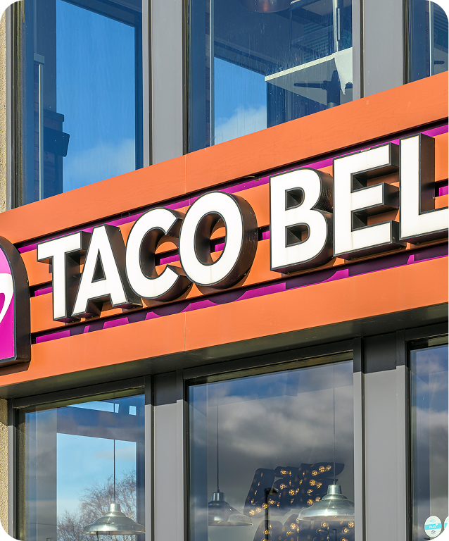 Close-up of a Taco Bell restaurant sign above glass windows, with reflections of the sky and nearby objects visible in the glass.