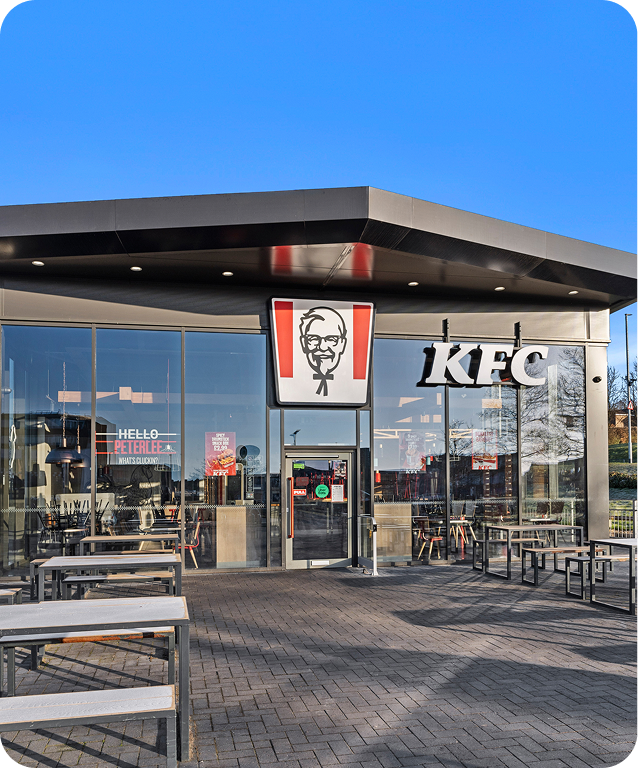 A modern KFC restaurant with large glass windows, outdoor seating, and the KFC logo above the entrance, under a clear blue sky.