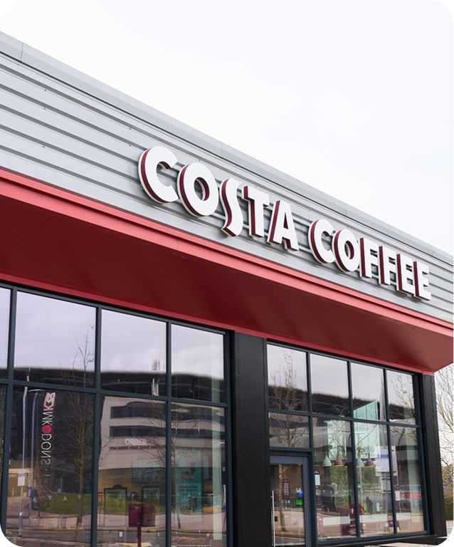 Storefront of a Costa Coffee café with large windows and a sign featuring the brand’s name in bold letters above the entrance. The building has a modern design with grey and red accents.
