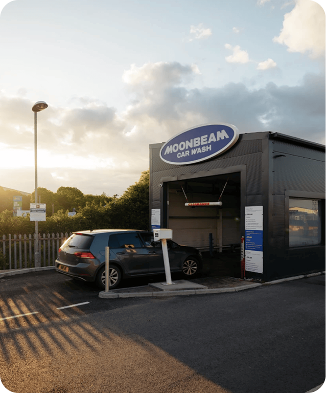 A dark-colored car is parked at the entrance of a Moonbeam Car Wash facility under a partly cloudy sky during sunset. The car wash building is gray with a rounded sign above the entrance.