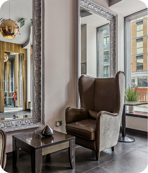 A modern lounge area with a brown leather wingback chair, a small wooden table, and a large ornate mirror on the wall, next to tall windows overlooking a city street.