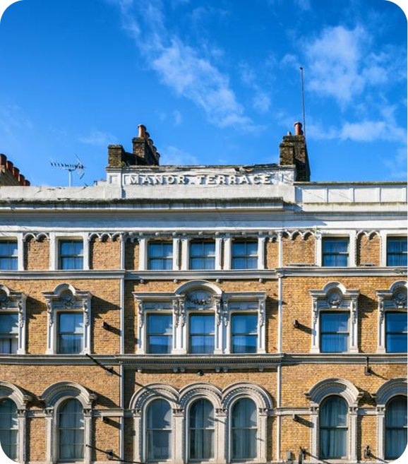 A historic brick building with arched windows and ornate stone details. The sign MANOR TERRACE is visible on the upper facade beneath a blue sky with scattered clouds.