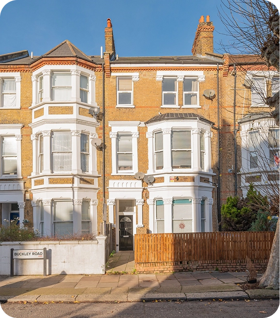 A three-story Victorian terraced house with bay windows, yellow brickwork, and white trim, located on Buckley Road. A wooden fence and a small garden are visible in front. Leafless tree branches frame the right side.