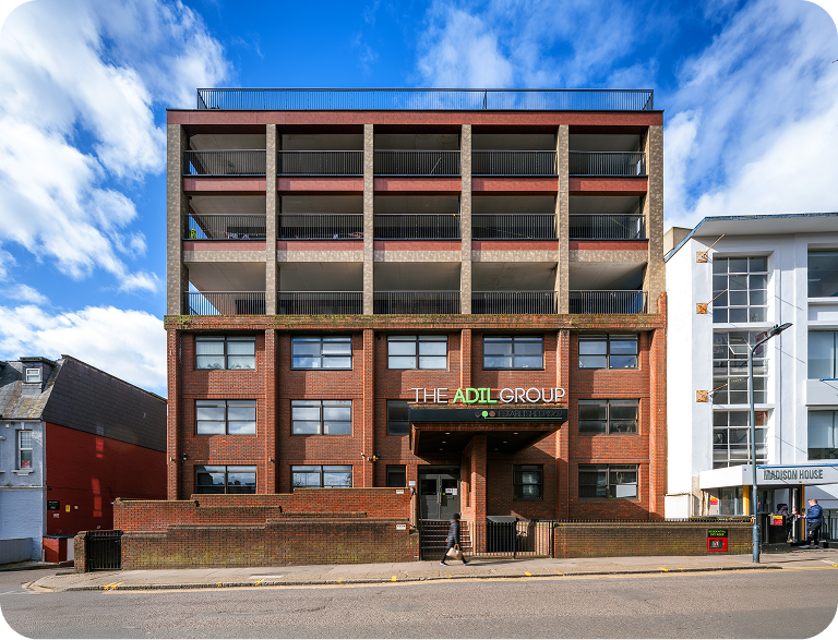A four-story brick office building with The Adil Group sign above the entrance, set against a partly cloudy sky. The building has balconies on the upper floors and is flanked by other modern structures.