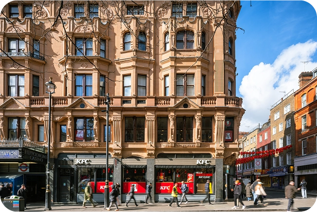 A busy city street with people walking past a historic building featuring a KFC restaurant on the ground floor, ornate architecture above, and a clear blue sky with some clouds.