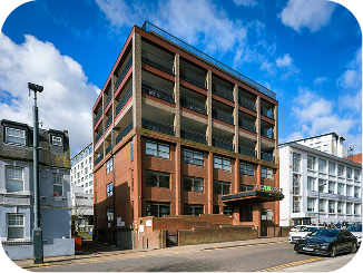 A red and brown multi-story office building with a flat roof stands on a city street under a blue sky with clouds. Cars are parked along the road, and adjacent white buildings are visible.