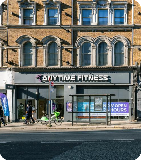 A street view of an Anytime Fitness gym in a historic brick building, with a bus stop, parked bicycle, and pedestrians passing by on a sunny day. Signs indicate the gym is open 24 hours.
