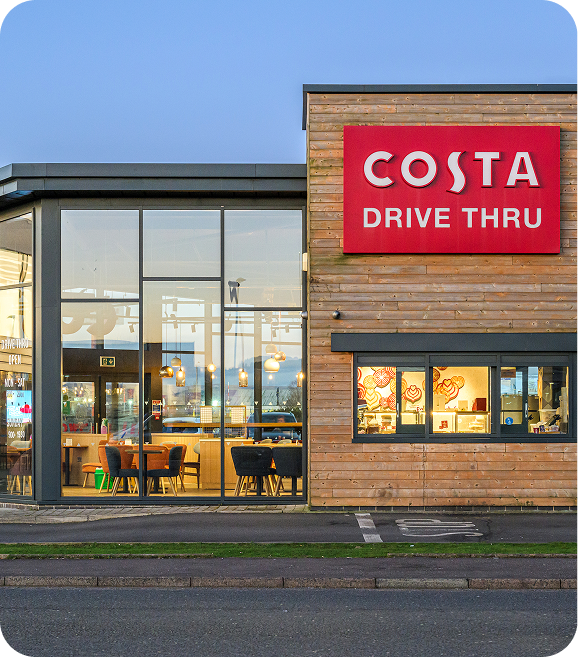 Modern Costa Coffee drive-thru with large glass windows, visible seating inside, and a prominent red Costa Drive Thru sign on the wooden exterior.