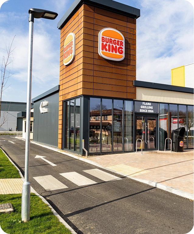 A modern Burger King restaurant with large windows and a tall sign on the front, located beside a paved road and sidewalk under a blue sky.