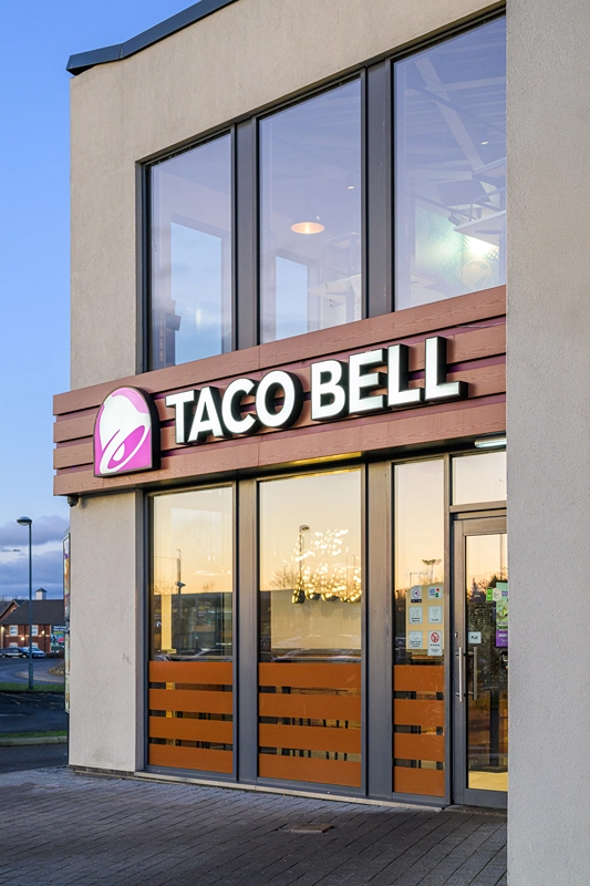 The exterior of a Taco Bell restaurant with large windows and a lit sign displaying the Taco Bell logo above the entrance. The sky and surrounding area are reflected in the glass.