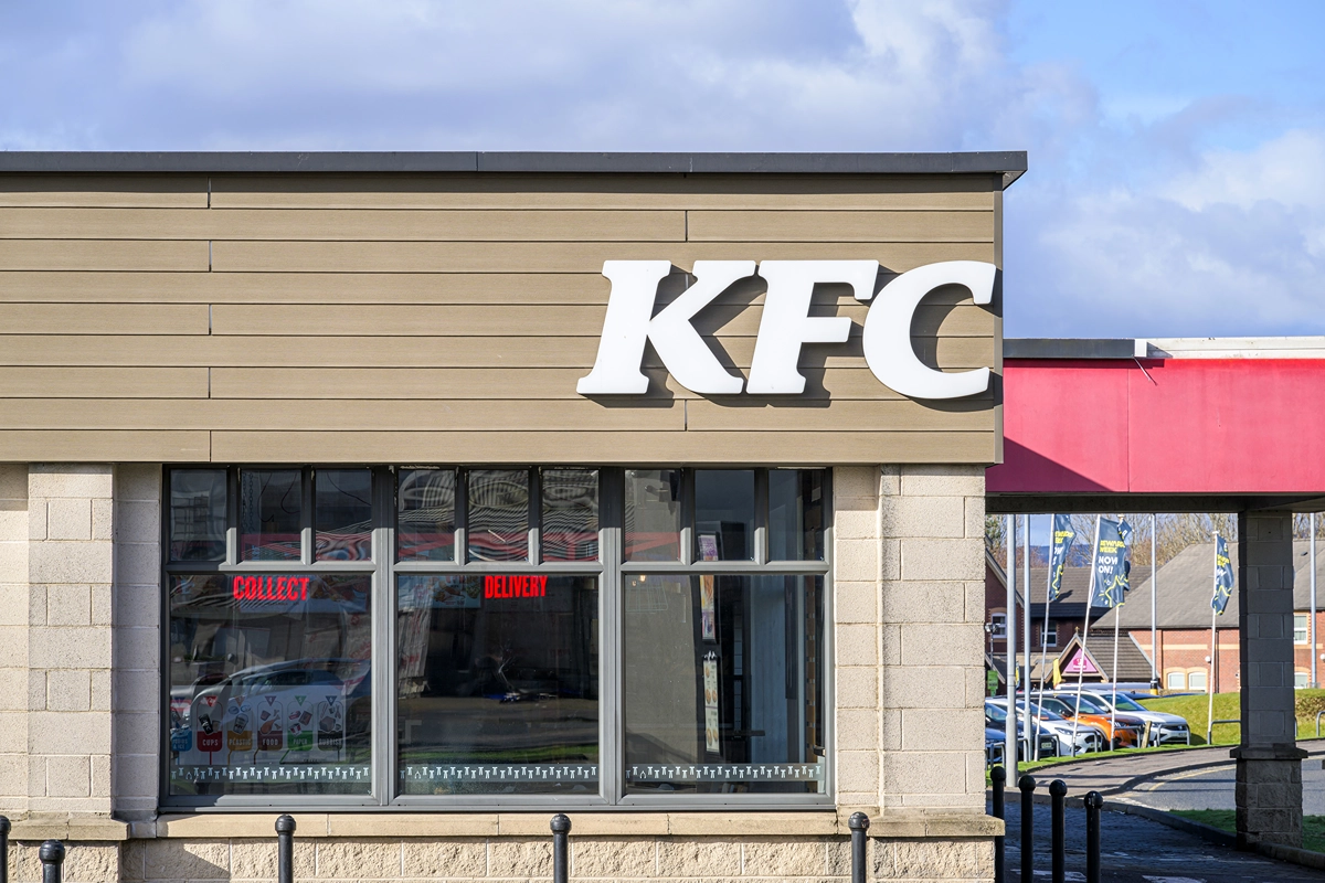 A KFC restaurant exterior with a large white KFC logo on a tan building above windows. Reflections and red signs reading “Collect” and “Delivery” are visible in the window. Cars and buildings are seen outside.