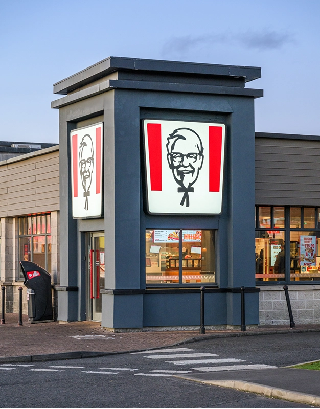 A KFC restaurant exterior featuring large illuminated signs with the KFC logo, showing Colonel Sanders’ face, above the entrance. The building is modern, and part of the drive-thru lane is visible in front.