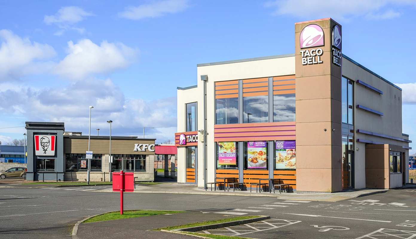 A Taco Bell restaurant with large windows and outdoor seating is in the foreground, while a KFC restaurant is visible in the background, both situated in a parking lot under a partly cloudy sky.