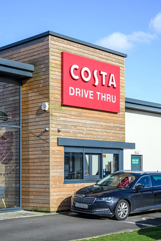 A black car parked at the drive-thru window of a modern Costa Coffee building, featuring a large red sign that reads COSTA DRIVE THRU on a wooden exterior wall.