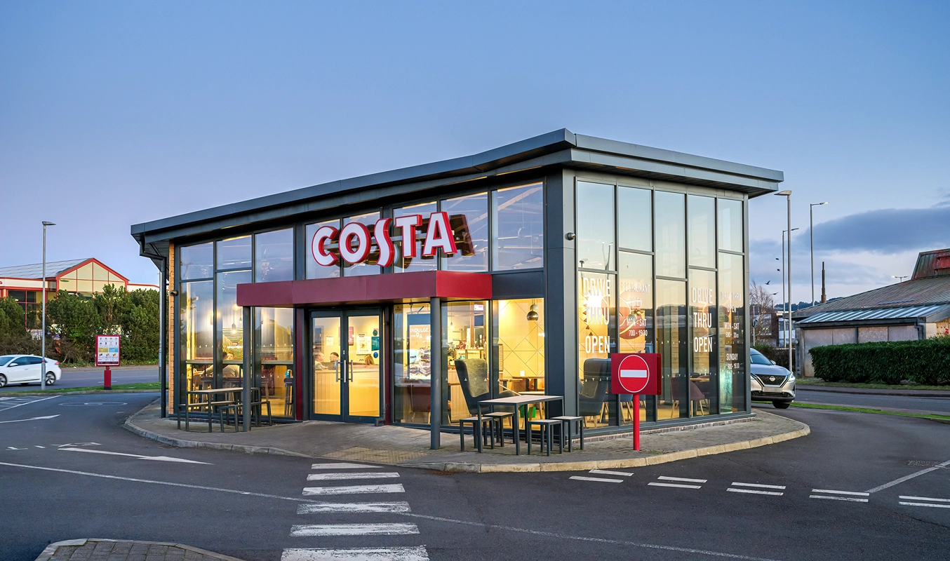 A modern Costa Coffee shop with large glass windows and a red sign, located on a corner with outdoor seating and a No Entry sign in the foreground. It is evening with lights on inside the café.