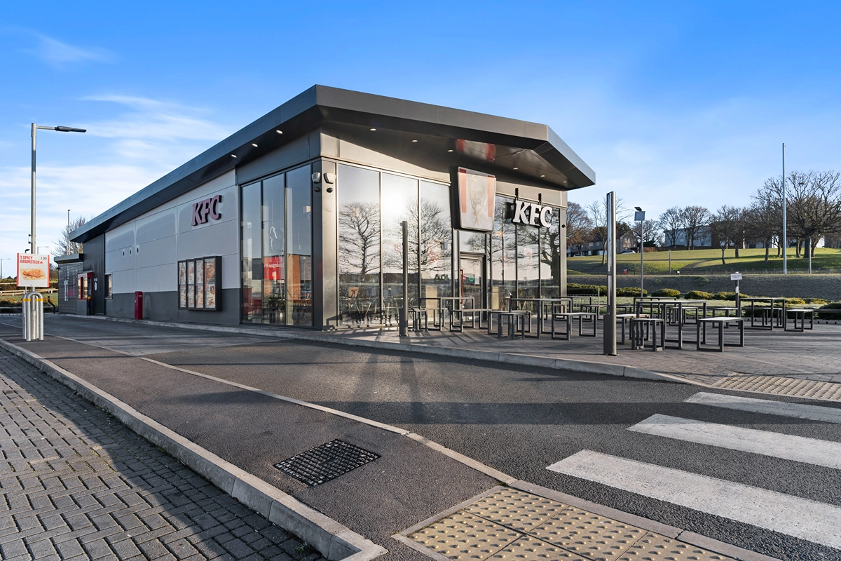 A modern KFC restaurant with large glass windows and outdoor seating is shown on a sunny day. The building is surrounded by pavement, a zebra crossing, and a grassy area with trees in the background.