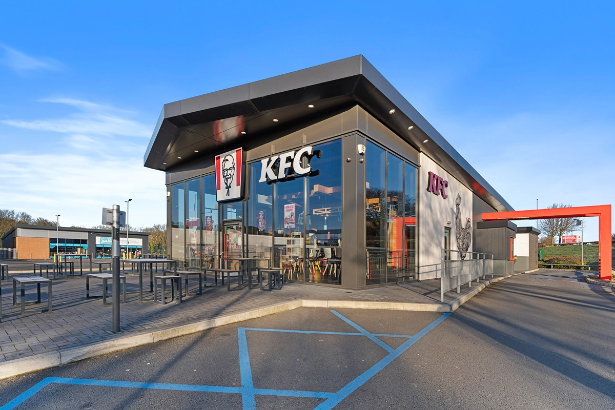 A modern KFC restaurant with large glass windows, outdoor seating, and drive-thru. The building displays the KFC logo and branding under a clear blue sky, with empty parking spaces in the foreground.
