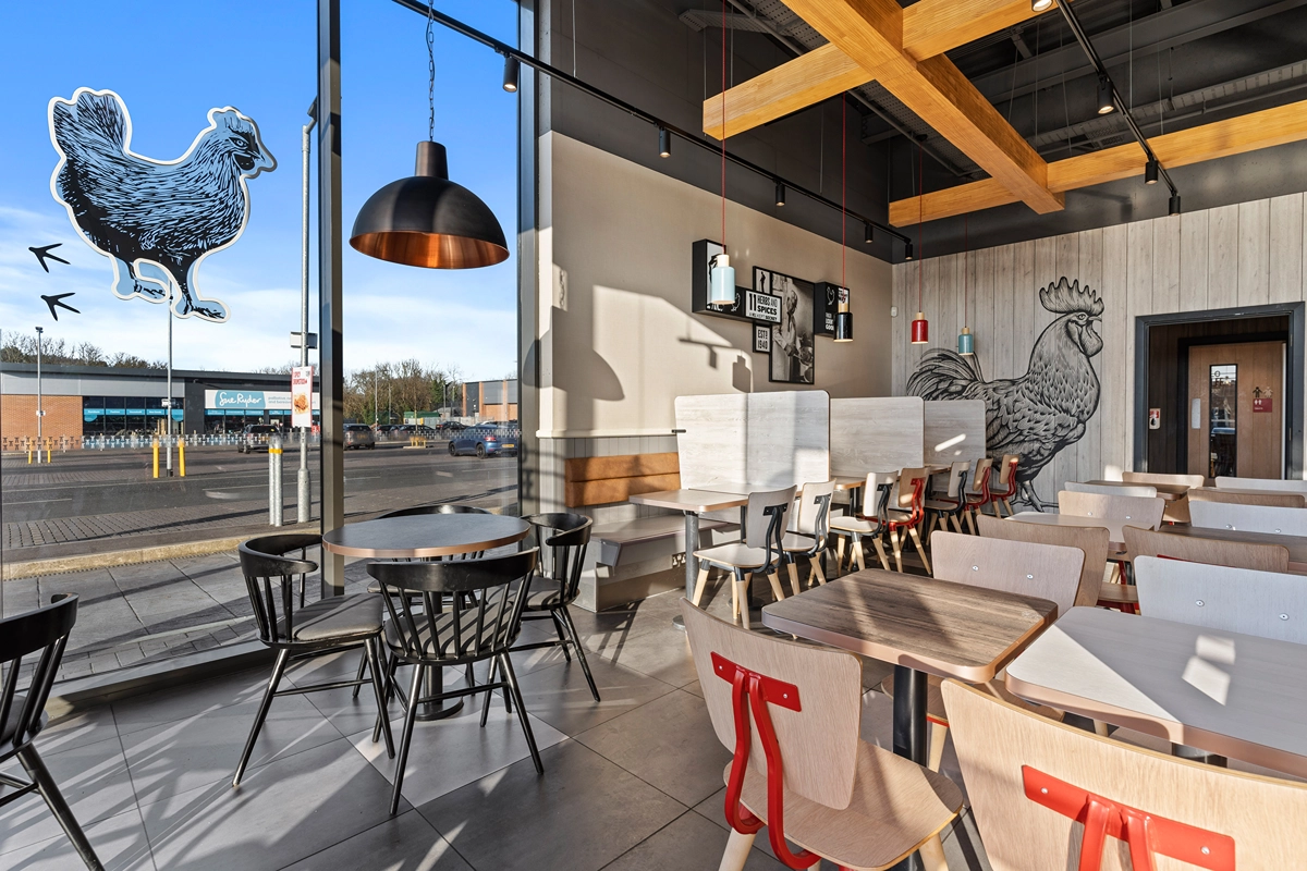 Modern fast-food restaurant interior with wooden tables and chairs, large windows, and rooster-themed wall art. Natural light fills the space, and a parking lot is visible outside through floor-to-ceiling windows.