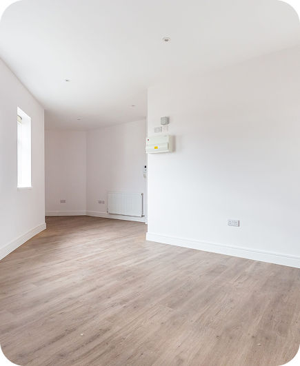 Bright, empty room with light wood flooring, white walls, a window on the left, and a radiator on the far wall. Electrical panels are mounted on the right wall; the ceiling has recessed lighting.