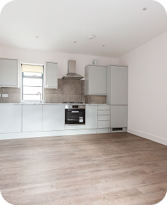 A modern, unfurnished kitchen with light grey cabinets, an oven, stovetop, and extractor hood. The room has light-colored walls, wood-style flooring, and a window letting in natural light.