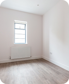 An empty, unfurnished room with light-colored walls, a single window, a radiator below the window, wood-style flooring, and baseboard trim along the walls.