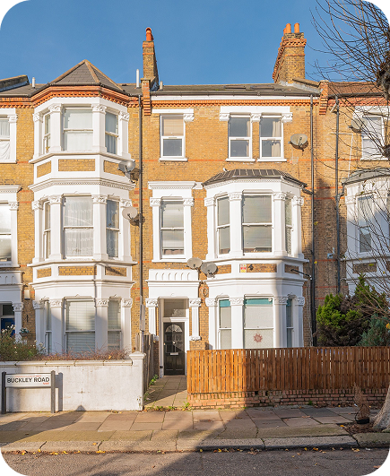 A row of three-story Victorian terraced houses with bay windows, white trim, and a small front yard with a wooden fence. A street sign reads Buckley Road. Trees without leaves line the sidewalk.