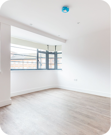 Bright, empty room with white walls, large windows letting in natural light, light wood flooring, and a blue smoke detector on the ceiling.