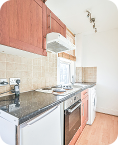 Modern kitchen with wooden cabinets, black countertop, electric stove, oven, washing machine, and beige tiled backsplash. Natural light enters through a window above the counter.