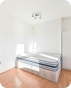 A small, bright bedroom with a striped mattress on a bed base, white walls, a wooden floor, a ceiling light fixture, and a closed white door. Sunlight streams through a window, creating a clean, minimal look.