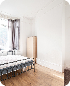 A bright, minimalist bedroom with a metal bed frame, mattress, light wood wardrobe, and sheer curtains on a window. The room has white walls and light wood flooring.