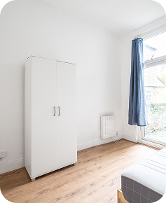 A bright, minimal bedroom with white walls, a white wardrobe, wood flooring, a radiator, a bed with a checkered duvet, and a large window with blue curtains letting in natural light.