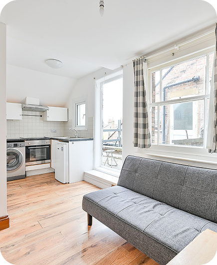 Bright, modern studio apartment with a small kitchen, washing machine, mini fridge, and oven. A gray sofa faces large windows with checkered curtains, letting in natural light. Light wood flooring throughout.