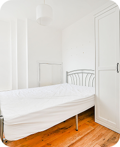 A small, bright bedroom with a metal bed frame, white bedding, a white wardrobe, wooden floor, and white walls, lit by natural light and a ceiling lamp.