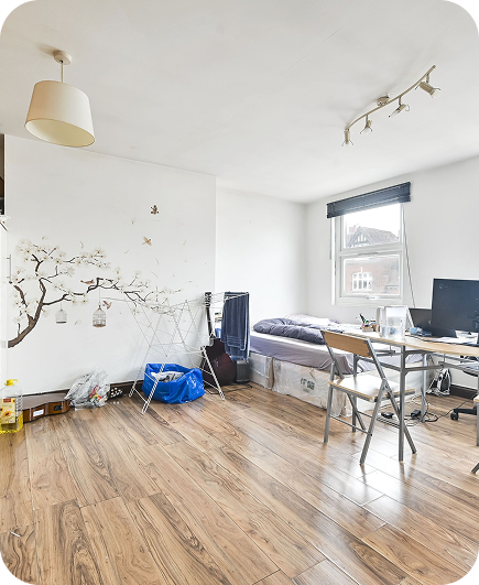 A bright, tidy bedroom with wood flooring, a single bed by the window, a desk with a computer, a chair, a drying rack, and a decorative tree mural on the white wall.