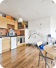 A small kitchen with light wood cabinets, a mini fridge, oven, and microwave. The floor is wooden, and a round table with metal chairs is in the foreground. A tree and bird wall decal decorates a white wall.