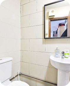 A small bathroom with white tiled walls, a white toilet, a pedestal sink with toiletries, and a rectangular mirror reflecting part of another room.