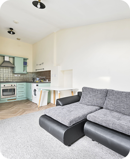 Modern, bright living space with a black and gray sectional sofa, light gray carpet, and a small kitchen area featuring mint green cabinets, an oven, a washing machine, and a dining table against a cream wall.