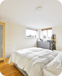 Bright bedroom with white walls, wood floor, a double bed with white bedding, a wooden wardrobe, a gray dresser under two windows, and a snowboard leaning against the dresser.