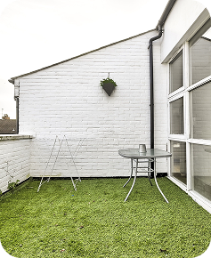 A small balcony with artificial grass flooring, a round glass table with two cups, a metal drying rack, and a hanging plant on a white brick wall next to large windows.