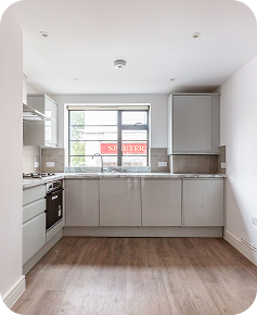 Modern kitchen with light grey cabinets, stainless steel appliances, a large window, and wood-style flooring. The space is clean, bright, and minimalistic with neutral tones throughout.
