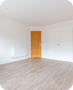 A bright, empty room with light-colored walls and wood flooring, featuring a closed wooden door, two white closet doors, and a white radiator on one wall.