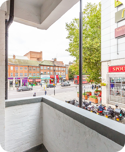 View from a small white painted balcony overlooking a busy street with parked scooters, pedestrians, shops, a McDonalds, a Sainsburys, and red double-decker buses in an urban area.