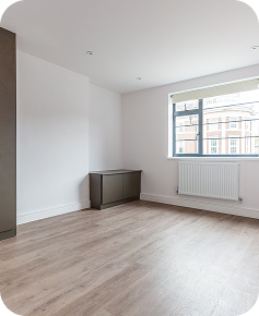 A bright, empty room with light wooden flooring, white walls, a large window with a radiator below it, and two gray cabinets along the walls.