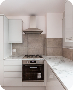 Modern kitchen with white cabinets, a stainless steel gas stove and oven, a vent hood, and marble-style countertops, featuring light-colored tiled backsplash and walls.