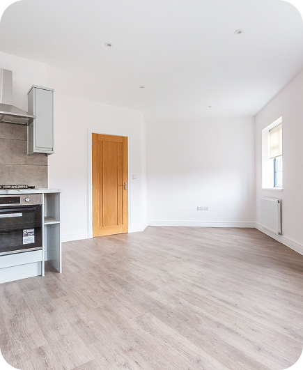 A modern, empty kitchen with light wood flooring, white walls, a wooden door, a window with a closed blind, and a built-in oven and stove on the left. The space is bright and minimalistic.