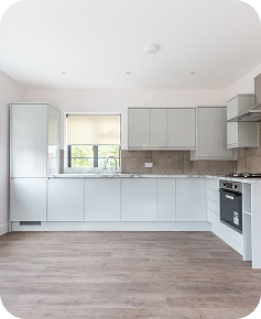Modern kitchen with light gray cabinets, built-in oven, gas stove, tiled backsplash, wood-style flooring, and a window letting in natural light. The space appears clean, bright, and minimalist.