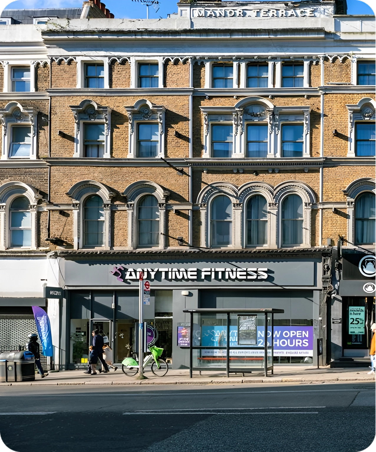 A brick building houses an Anytime Fitness gym on the ground floor. Two people walk past, and a green bicycle is parked outside. Signs on the gym windows announce it is now open 24 hours.