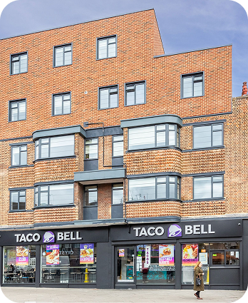 A Taco Bell restaurant on the ground floor of a modern brick building with large windows. A person is walking past the storefront, and colorful posters are visible in the windows.