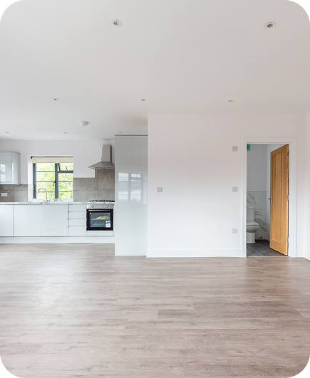 A modern, unfurnished kitchen and living area with white walls, light wood flooring, stainless steel appliances, and a wooden door leading to a bathroom. Natural light comes in through the window.