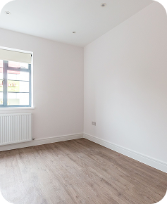 A small, empty room with white walls, a wooden floor, a window with a shade partially down, and a white radiator underneath the window. The room is well-lit with natural light.